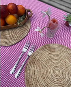 Dinner table setting. pink/ cerise Gingham table cloth with Palm leaf placemats on top decorates with forks amd a bowl full of blood oranges 